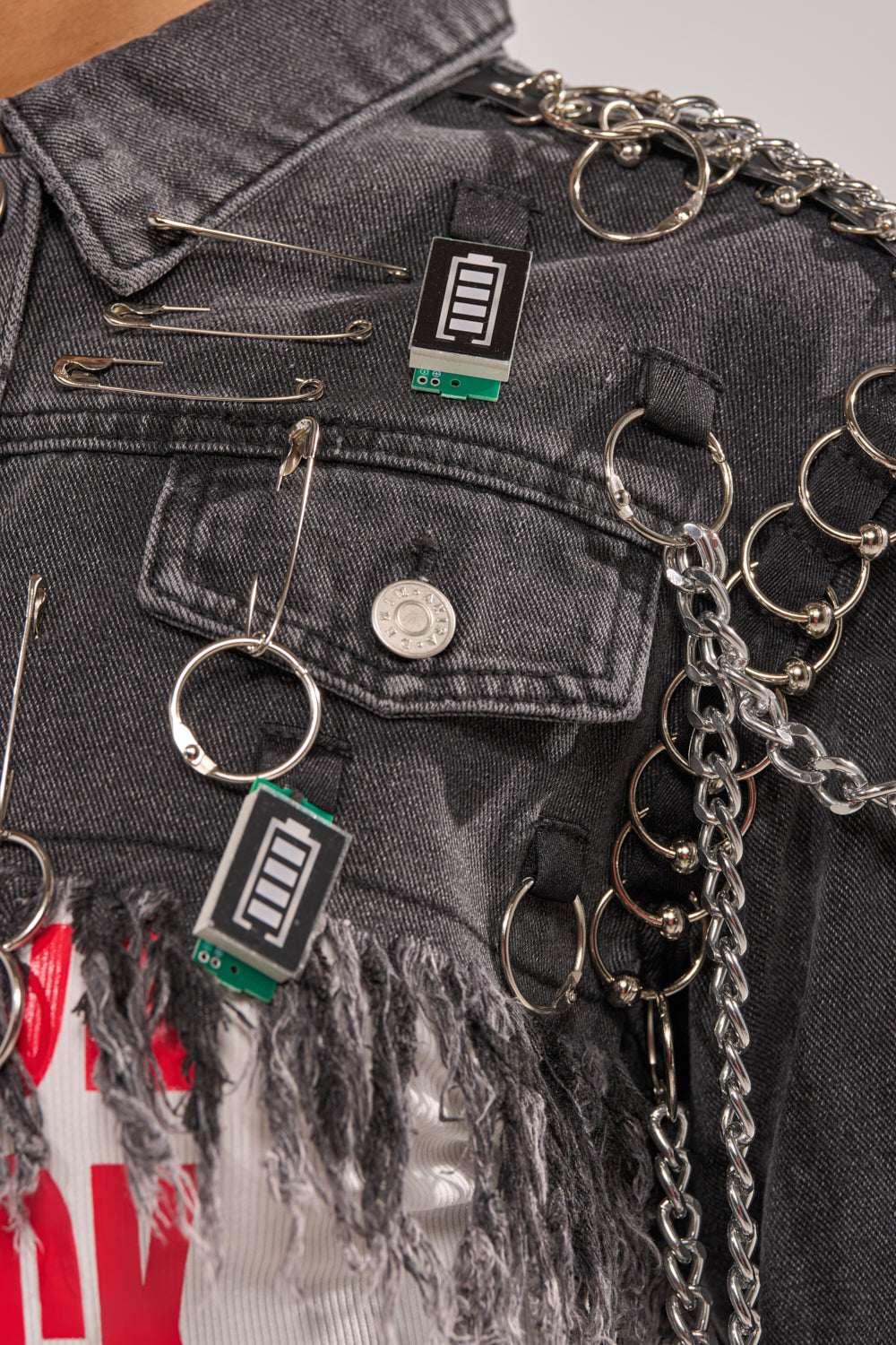 Close-up of the CAN'T COMPUTE ULTRA CROPPED DENIM JACKET, detailed with safety pins, metal chains, rings, fringe, and small electronic circuit boards. A white shirt with red print is visible under the distressed hem. 5