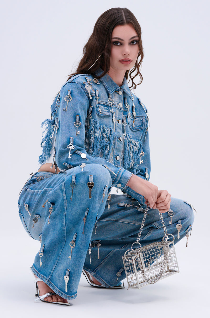 Wearing the KEY TO THE CITY CROPPED DENIM JACKET and matching jeans adorned with silver keys, a woman poses confidently. She holds a silver wire handbag, wears silver heels, and lets her wavy hair flow freely against a simple background.
