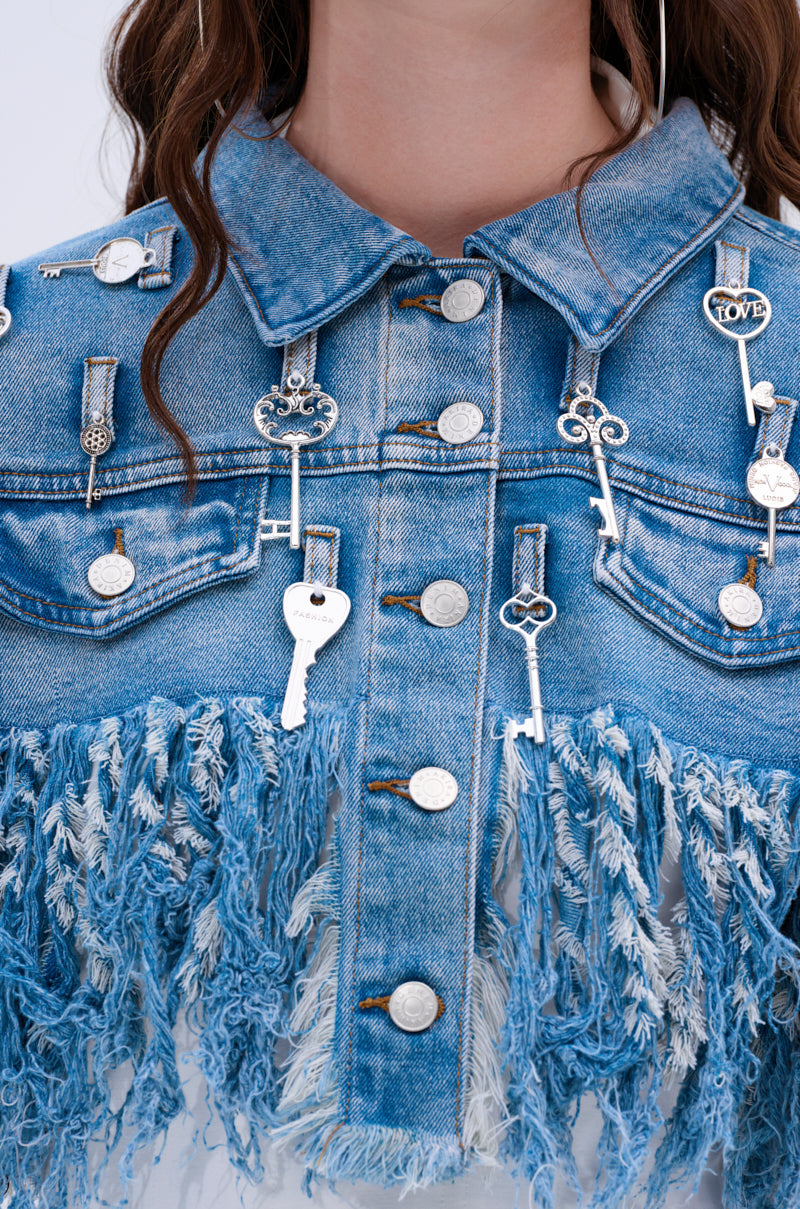 Close-up of the KEY TO THE CITY CROPPED DENIM JACKET, highlighting its distressed fringe, silver key and charm pins, collar, buttons, and upper chest area.