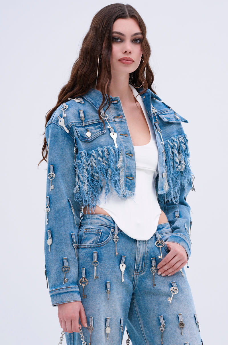 A woman with long wavy brown hair wears the KEY TO THE CITY CROPPED DENIM JACKET and jeans featuring silver keys and distressed fringe over a white fitted top, accessorized with large hoop earrings against a plain light background.