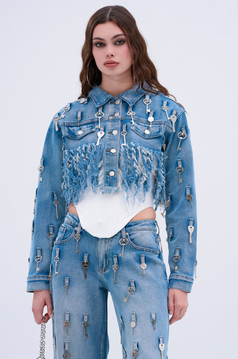 Wearing the KEY TO THE CITY CROPPED DENIM JACKET with frayed chest detailing, a woman pairs it with jeans adorned in silver keys. She stands against a plain light background, holding a small chain strap purse.