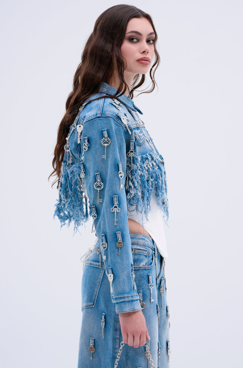A woman with long wavy brown hair wears the KEY TO THE CITY CROPPED DENIM JACKET with a matching skirt featuring distressed fringe and chains, posing sideways against a plain light background.