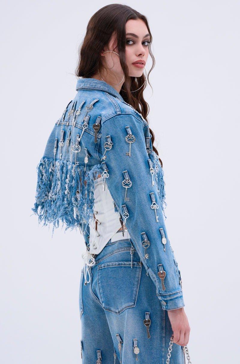 Wearing the KEY TO THE CITY CROPPED DENIM JACKET with distressed fringe and silver key details, a woman with long wavy hair looks over her shoulder against a plain light background.