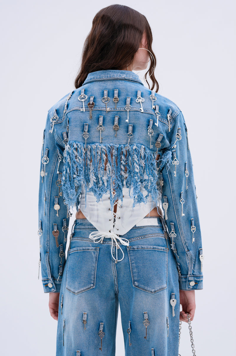 A person with long wavy hair, seen from behind, wears the KEY TO THE CITY CROPPED DENIM JACKET laced at the back, paired with jeans featuring silver key accents and distressed fringe for an edgy style.