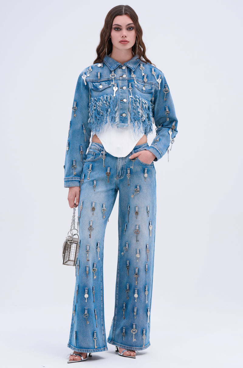 A woman wears the KEY TO THE CITY CROPPED DENIM JACKET and matching jeans with metallic key-shaped embellishments, holding a small silver bag against a plain light background.