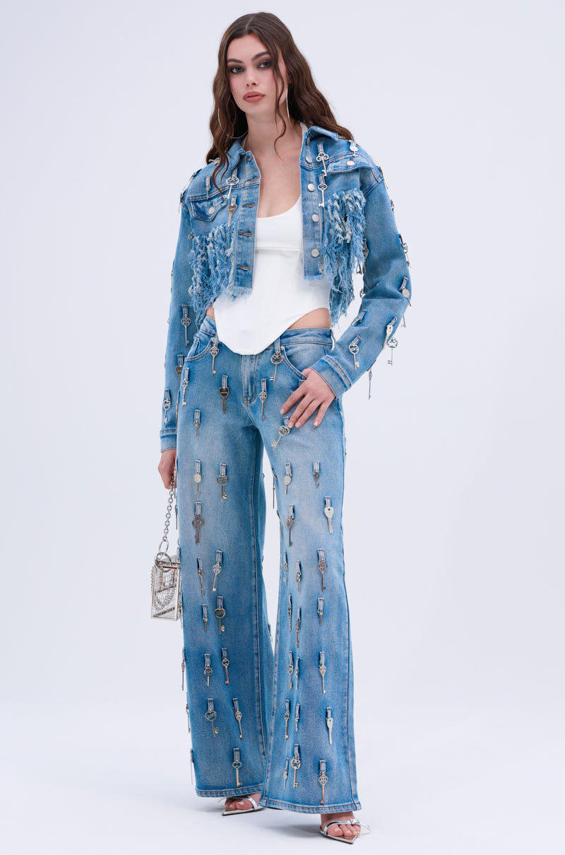 A woman wears the KEY TO THE CITY CROPPED DENIM JACKET and matching jeans with silver keys, paired with a white top and metallic purse, standing against a plain light background.
