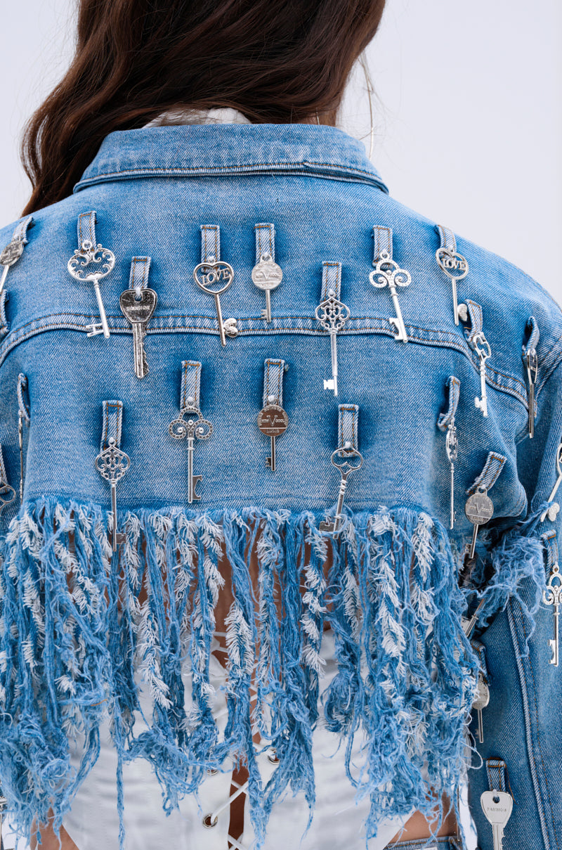 A person wears the KEY TO THE CITY CROPPED DENIM JACKET, featuring silver keys on the back and distressed fringe trim. The background is a plain light color.