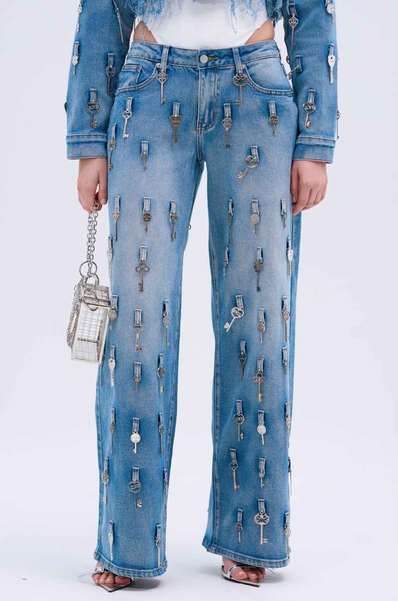 Wearing KEY TO THE CITY RELAXED FIT JEANS—inspired blue, mid-rise denim with metal key details—a person holds a small metallic chain purse and wears open-toed heels, all set against a plain white background.