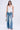 A woman stands against a plain background wearing a white halter top, KEY TO THE CITY RELAXED FIT JEANS with a mid-rise and metal key details, silver high-heeled sandals, and holding a small silver handbag.
