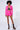 A person stands against a plain background wearing a vibrant pink cropped jacket with matching Better Luck Utility Pocket Shorts in Pink, a black bralette, calf-high black socks, and leopard-print platform heels. They have a curly afro hairstyle and a confident expression.