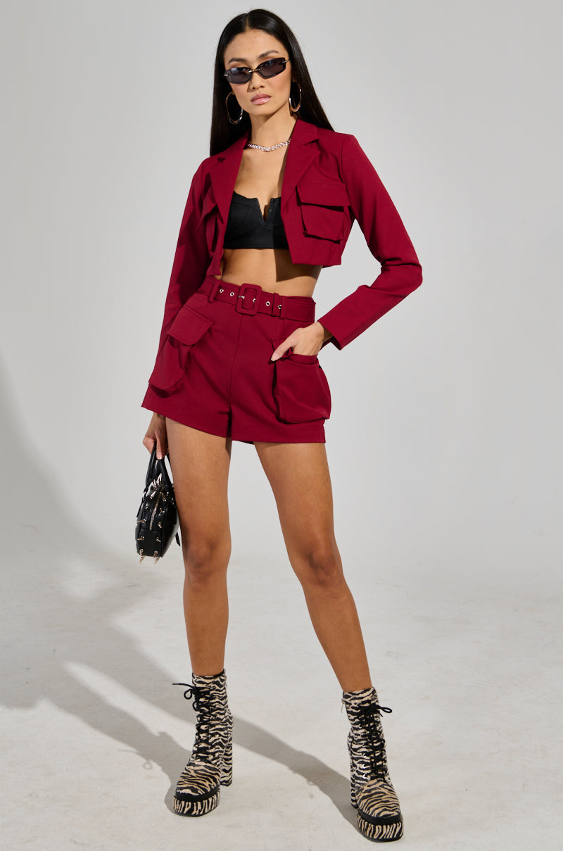 Dressed in the chic BETTER LUCK UTILITY POCKET SHORTS IN BURGUNDY, a person showcases their style by pairing them with a matching jacket, a sleek black crop top, and eye-catching zebra-patterned boots. They complete the look with sunglasses, jewelry, and a stylish black handbag while striking a confident pose against a plain background.