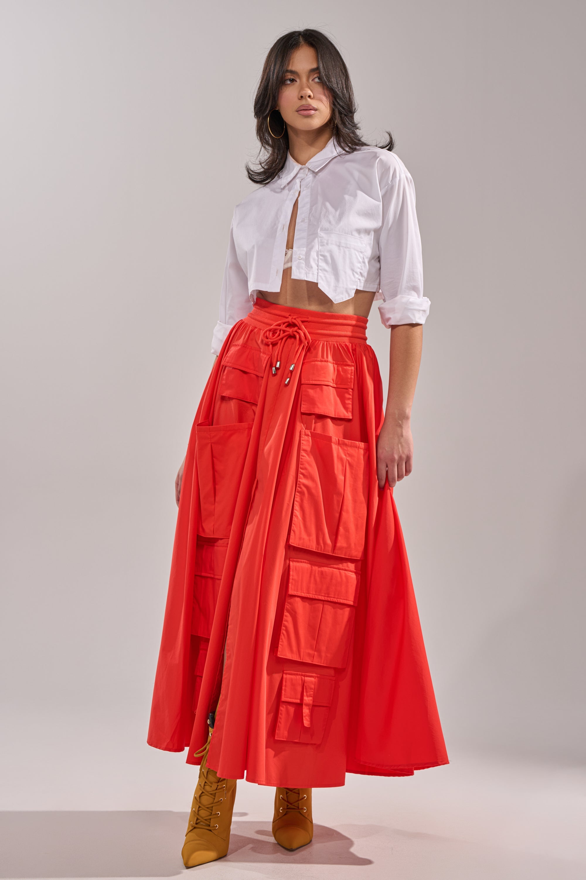 A woman poses in a white cropped button-up shirt and the AMELIA CARGO POCKET MAXI SKIRT IN RED, which features multiple large pockets. She completes the look with mustard yellow lace-up boots against a neutral background. 0
