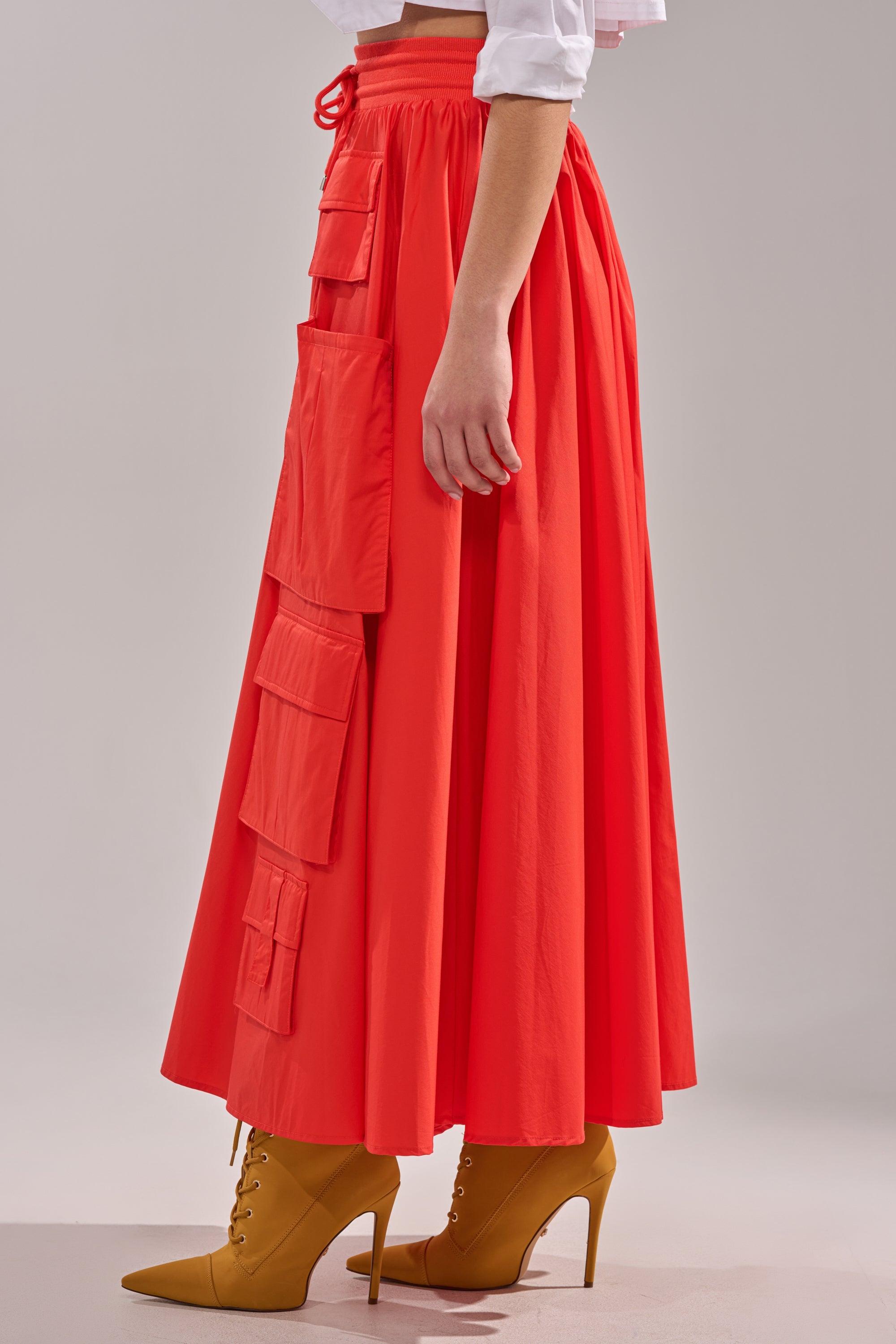 A person wears the AMELIA CARGO POCKET MAXI SKIRT IN RED with large square pockets, styled with mustard yellow heeled lace-up boots against a plain light background. 3