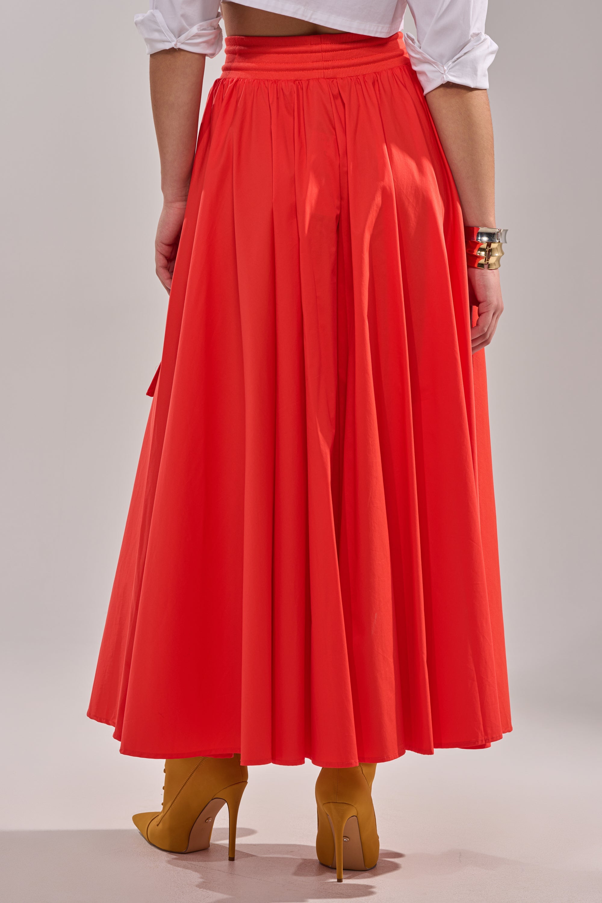 A person seen from the back wears the AMELIA CARGO POCKET MAXI SKIRT IN RED with a white top, mustard yellow high-heeled boots, and gold bracelets. 4