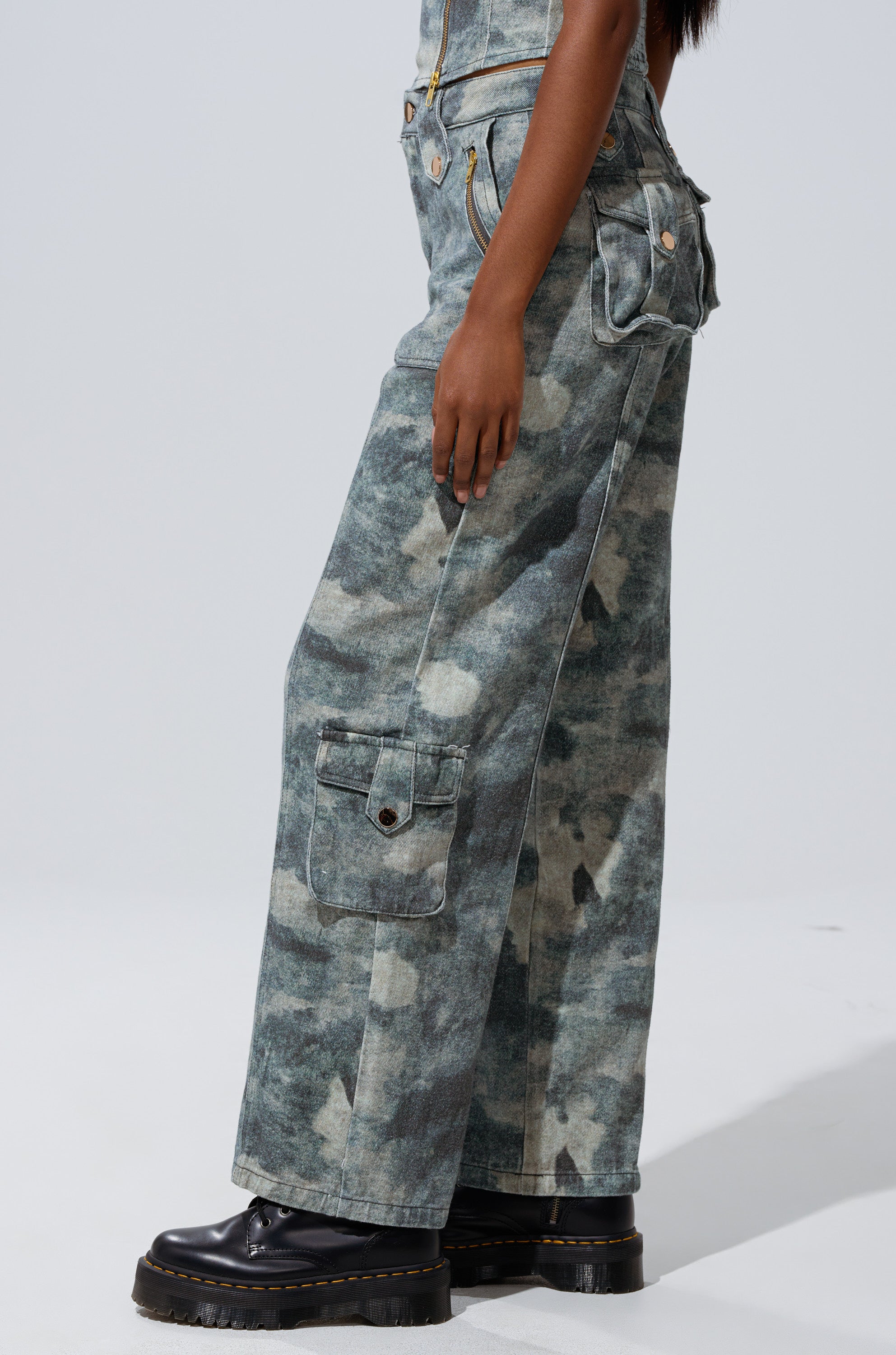 The lower half of a person is shown wearing STEPH CAMO DENIM RELAXED PANT, featuring loose, wide-leg cargo style with flap pockets, paired with black platform shoes. The backdrop is plain and light, highlighting street-style flair. 3