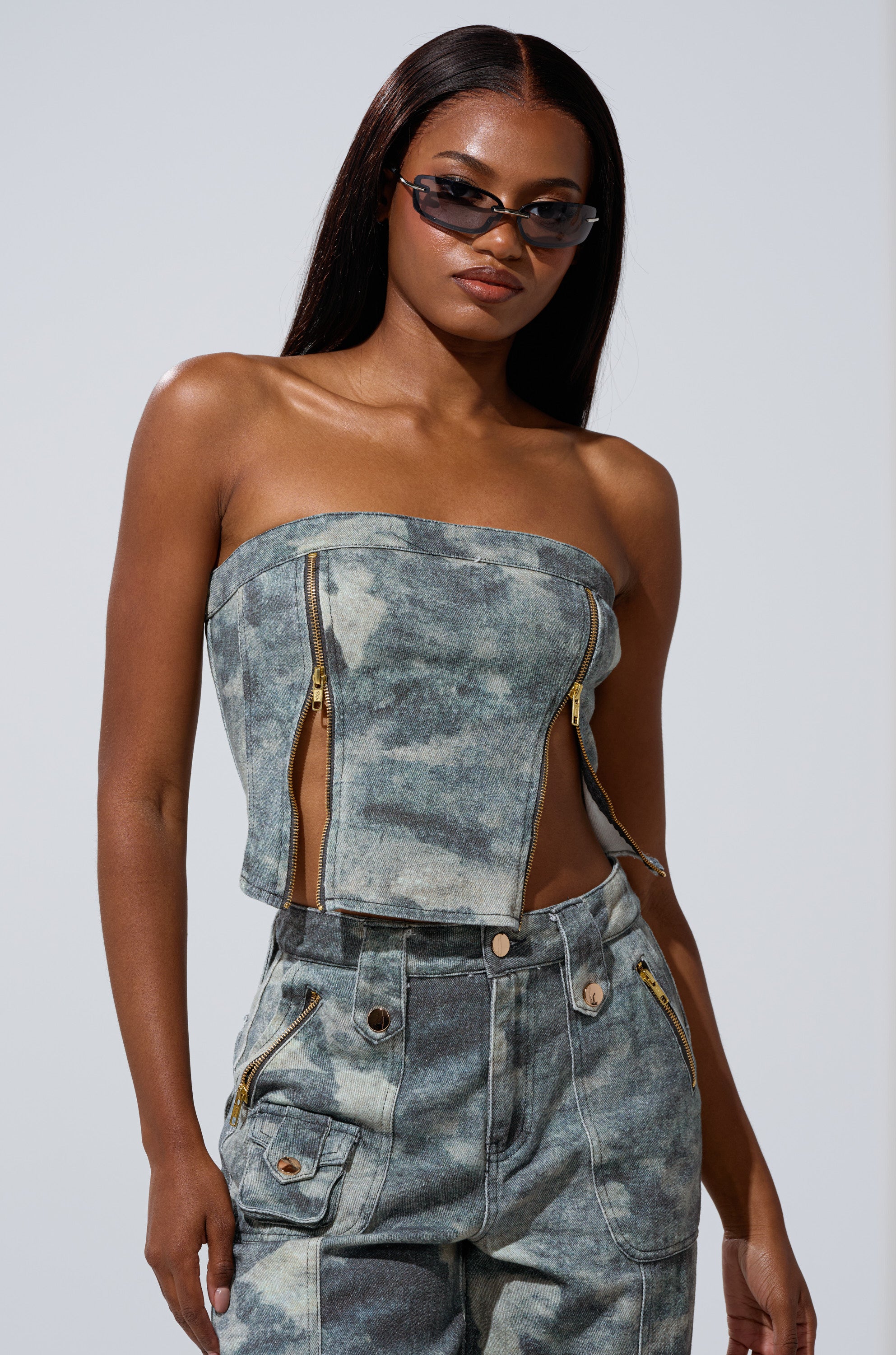 Wearing the STEPH CAMO DENIM CORSET with front zippers and matching high-waisted pants, a woman channels utilitarian chic, standing confidently with long straight hair and narrow sunglasses.