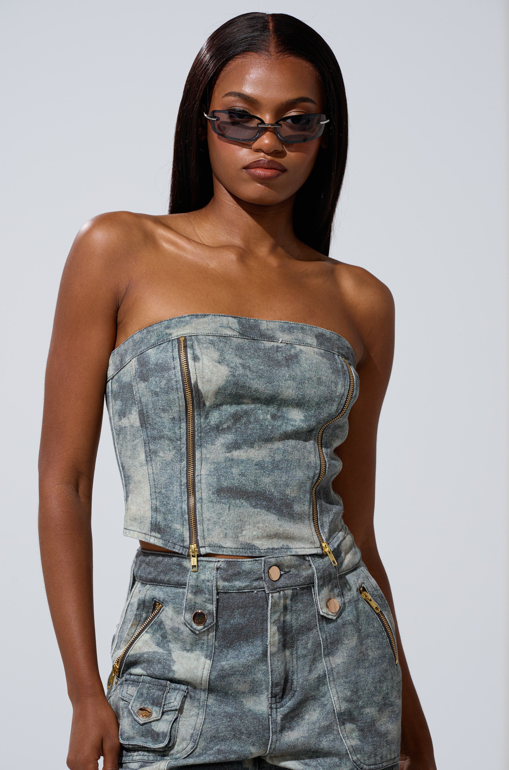 Wearing the STEPH CAMO DENIM CORSET and matching high-waisted shorts with zippers and pockets, she exudes utilitarian-chic style, accessorized with narrow black sunglasses as she poses confidently against a plain background.