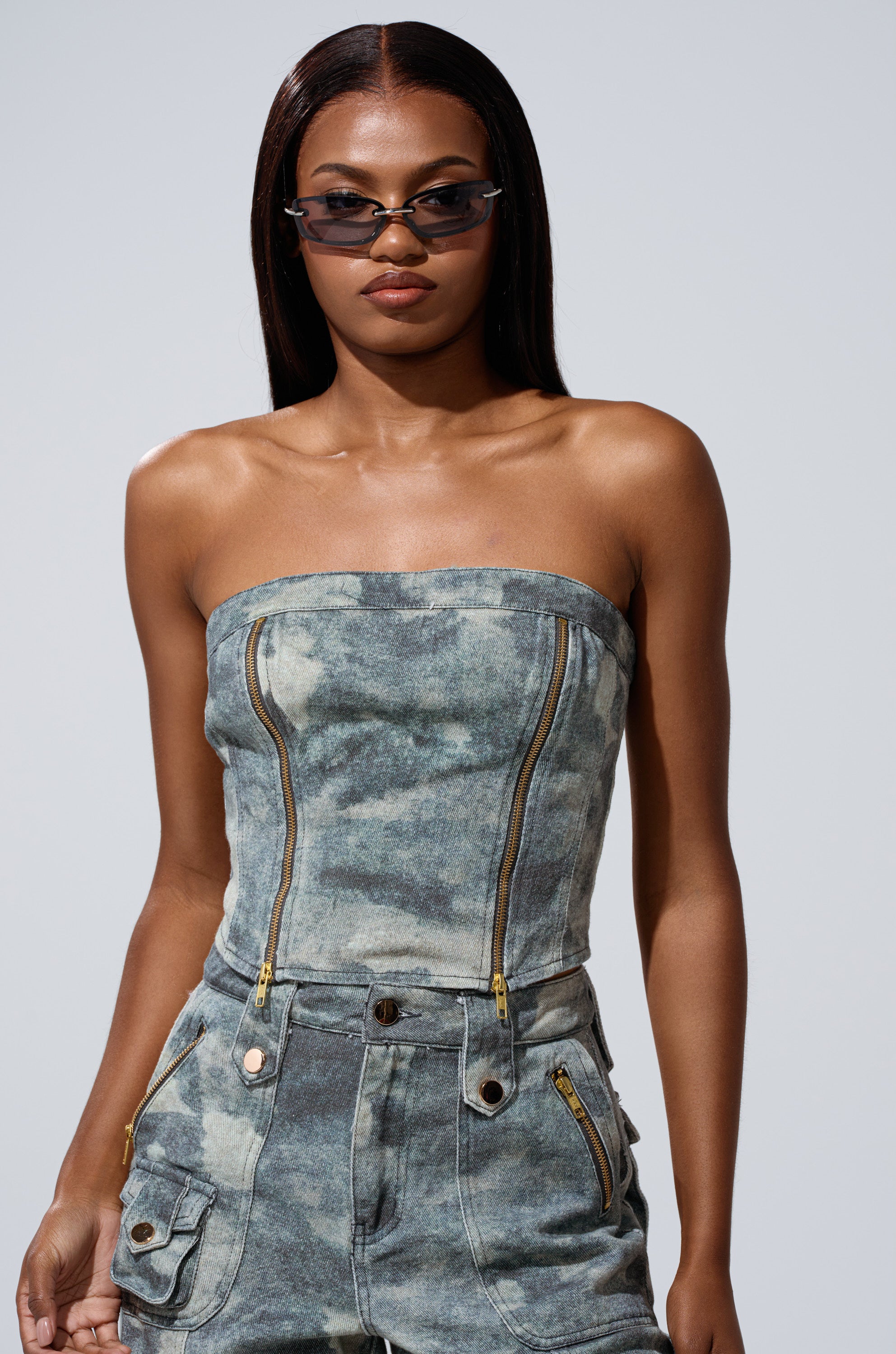 A woman in the STEPH CAMO DENIM CORSET and matching high-waisted pants, styled with sunglasses for a utilitarian chic look, stands against a plain background.