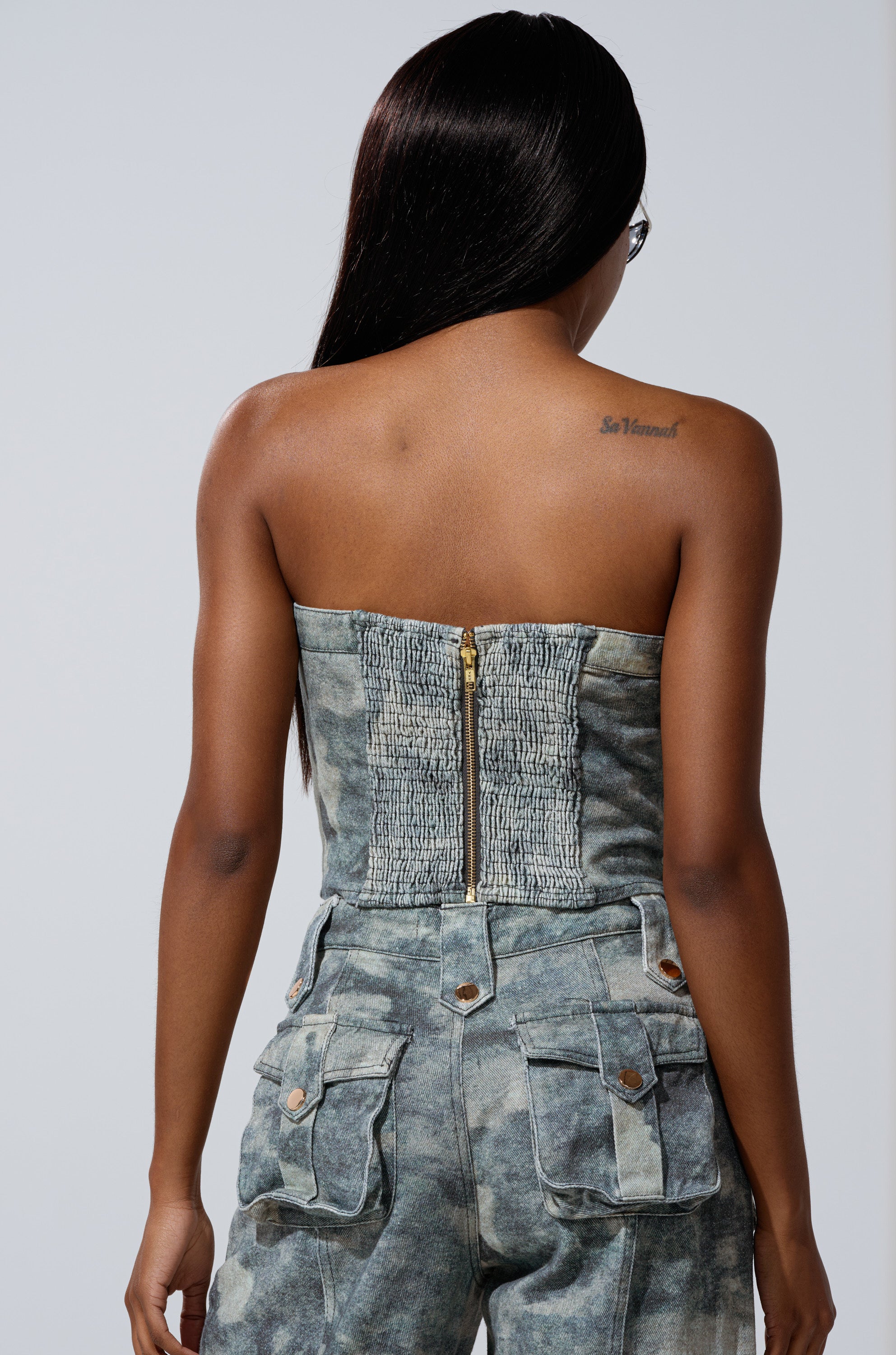 A woman with long, straight dark hair faces away, wearing the STEPH CAMO DENIM CORSET with a visible zipper and matching high-waisted pants. Her upper back tattoo reads "So Loved” for a bold utilitarian-chic look.