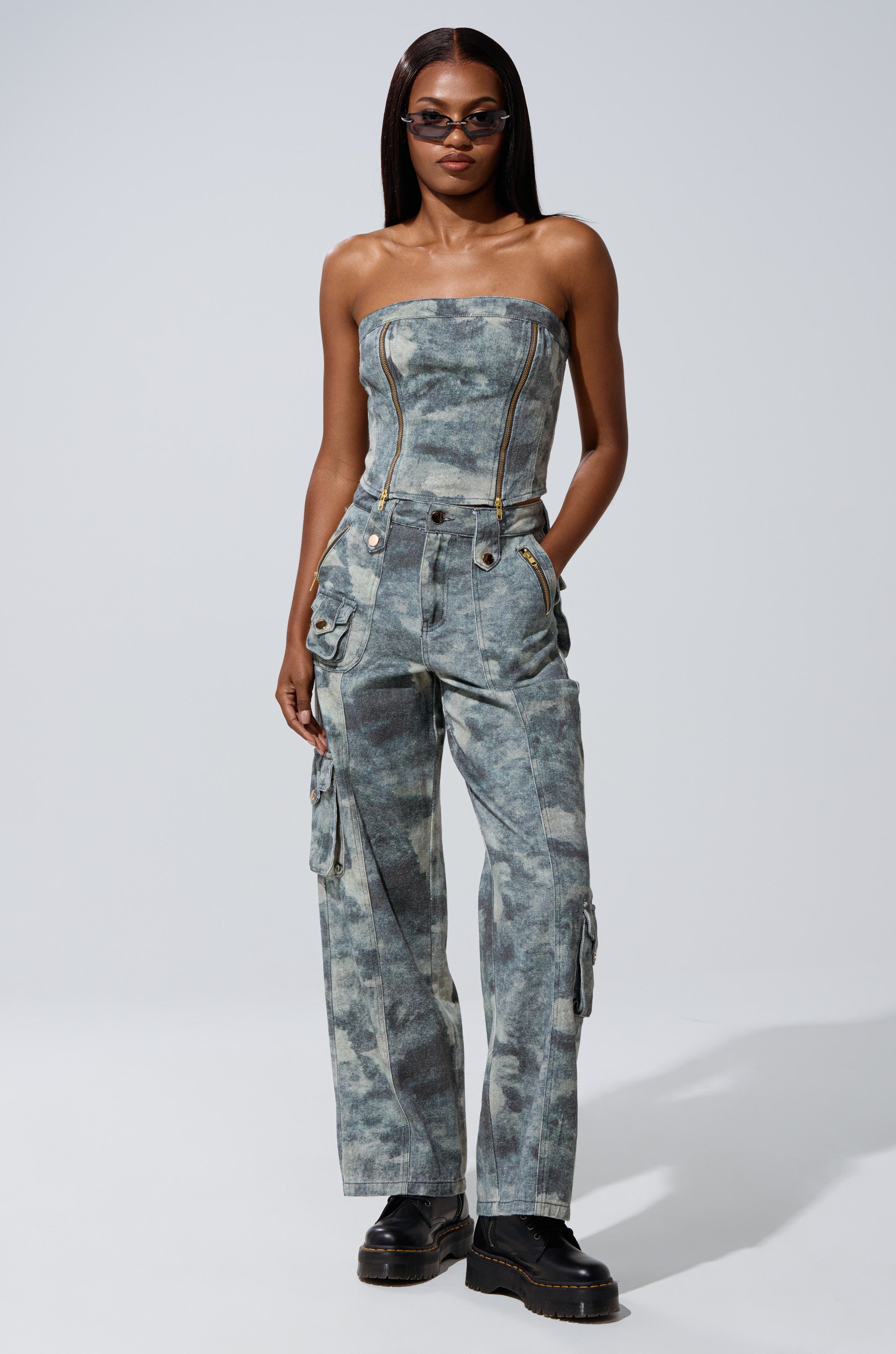Wearing the STEPH CAMO DENIM CORSET and matching cargo pants with zippers and pockets, she channels utilitarian chic. With straight hair, sunglasses, and black platform shoes, she stands confidently with hands in her pockets.