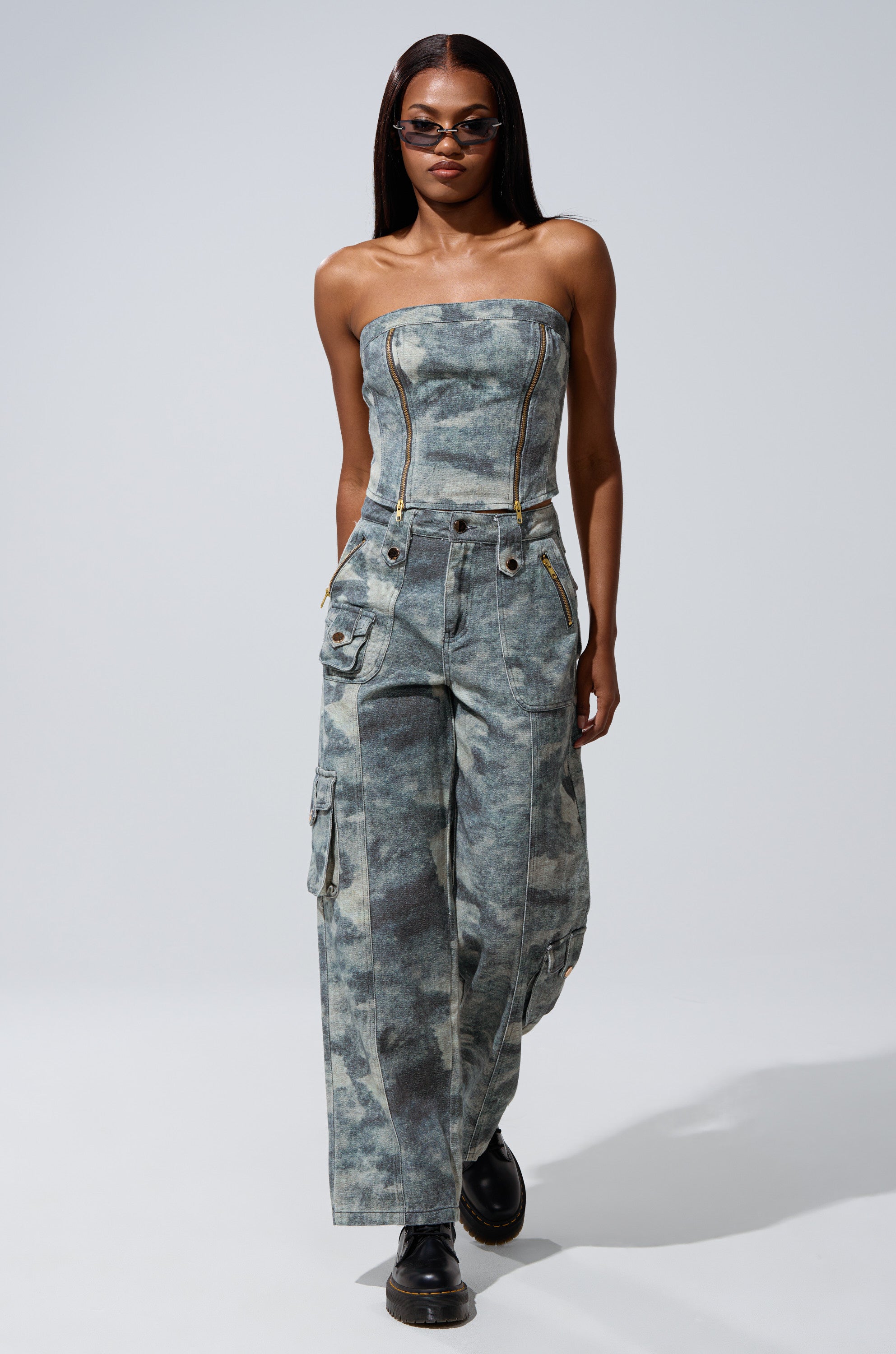 A woman exudes confidence in the STEPH CAMO DENIM CORSET, styled with straight hair, sunglasses, and black platform shoes. The utilitarian-chic piece features a strapless silhouette with cargo pockets and zipper accents against a plain backdrop.