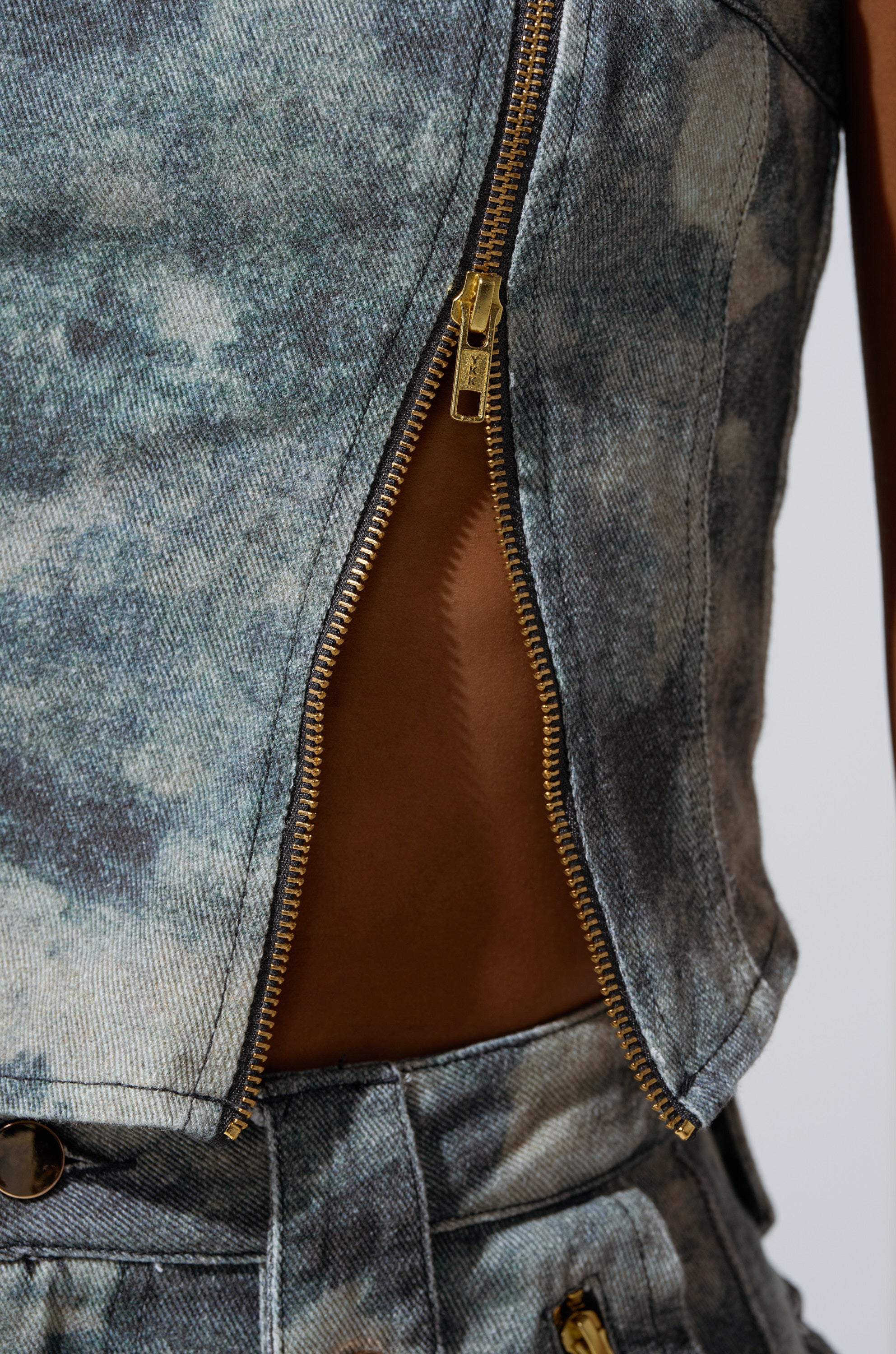 A close-up of the STEPH CAMO DENIM CORSET shows its blue, acid-washed denim and a side gold zipper slightly unzipped, blending utilitarian chic with a strapless design that reveals skin.