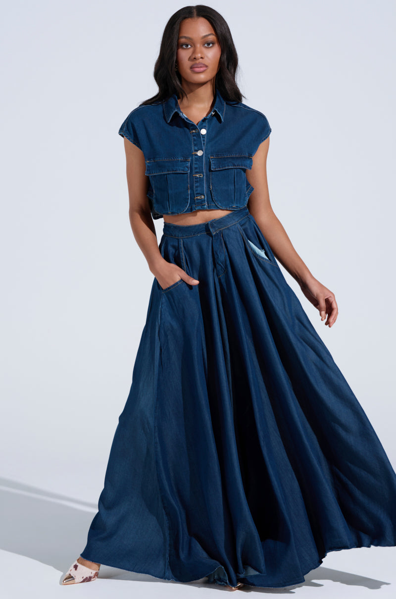 A woman dons an AKIRA Label blue denim cropped vest and a CARRIE ULTRA WIDE LEG DENIM TROUSER, confidently posing with her hand in her pocket against a minimalistic background. The high-rise design combines style with ease.