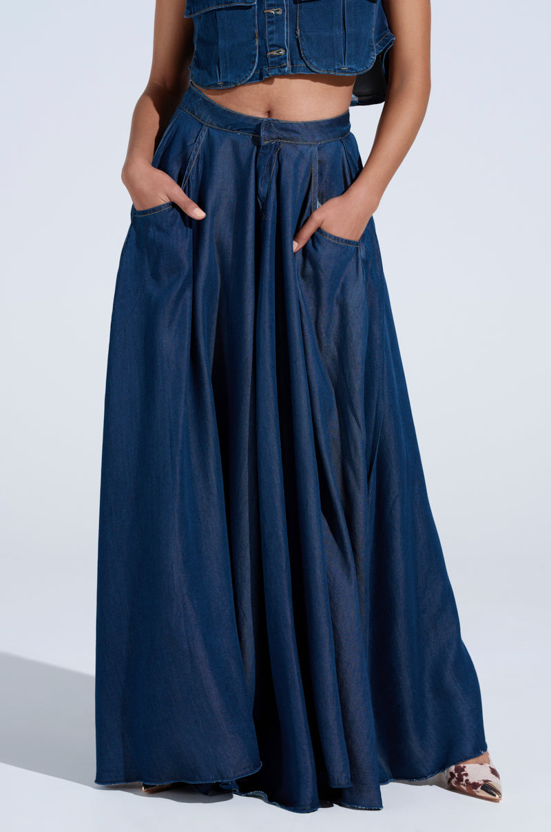 Styled in a long, flowing dark denim skirt with hands in pockets, they exude an AKIRA Label vibe. Paired with a matching denim top against a plain background, they embrace the charm of CARRIE ULTRA WIDE LEG DENIM TROUSER aesthetics.