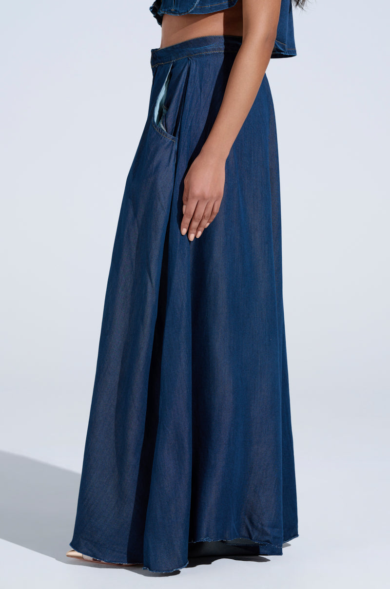 Someone is presenting the AKIRA Label ensemble with the CARRIE ULTRA WIDE LEG DENIM TROUSER, a long dark blue denim skirt featuring wide-leg elegance and practical pockets, standing sideways against a plain, light background.