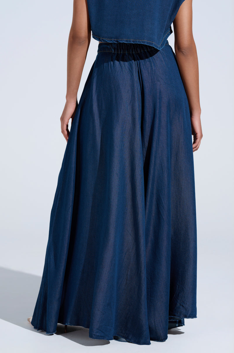 Against a pale background, a person wears the CARRIE ULTRA WIDE LEG DENIM TROUSER with an elastic waistband. The dark wash denim skirt mimics the fluid flow of wide-leg trousers, moving elegantly with each slight motion as their relaxed arm hangs by their side.