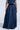Against a pale background, a person wears the CARRIE ULTRA WIDE LEG DENIM TROUSER with an elastic waistband. The dark wash denim skirt mimics the fluid flow of wide-leg trousers, moving elegantly with each slight motion as their relaxed arm hangs by their side.