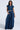 A woman poses against a white background, wearing a sleeveless denim crop top and the CARRIE ULTRA WIDE LEG DENIM TROUSER, featuring high-rise, dark wash denim with pockets. One hand rests in her pocket while the other hangs gracefully by her side.