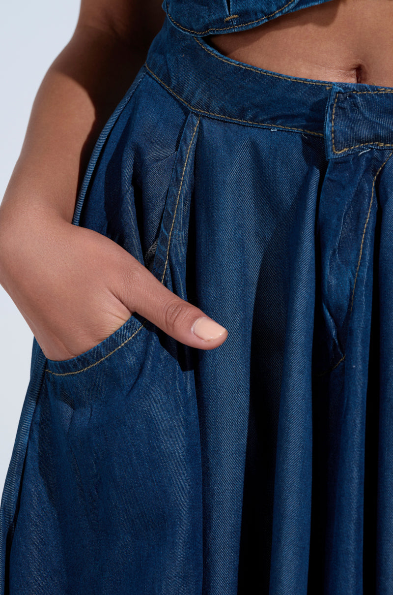 A close-up shows a hand in the pocket of the CARRIE ULTRA WIDE LEG DENIM TROUSER. The dark wash denim features visible stitching and pleats, with medium skin tone highlighting its rich texture and intricate design.