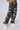 A person stands with one hand on their hip, wearing the APPLY PRESSURE GREY CAMO CARGO PANT from AKIRA Label, adorned with black beads and paired with white sneakers. The plain background enhances the focus on the utility-inspired fashion.