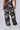 A person is standing with their back to the camera, wearing an AKIRA Label APPLY PRESSURE GREY CAMO CARGO PANT adorned with black beads. The outfit features utility-inspired cargo pants paired with light-colored sneakers against a plain white background.