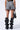 A person stands facing away from the camera, wearing the MIDDAY PARK DATE HIGH WAIST SHORT in gray acid wash denim with frayed hems, a black crop top, and black high-heeled boots featuring wave-like designs and studs. They carry a black handbag with eyelet details.