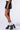 A person is seen wearing the LUCKY LOO MINI SKIRT, a black faux leather mini skirt featuring a pleated front design, along with white socks and black high-heeled sandals adorned with silver studs. The individual is photographed from the thigh down against a light background.