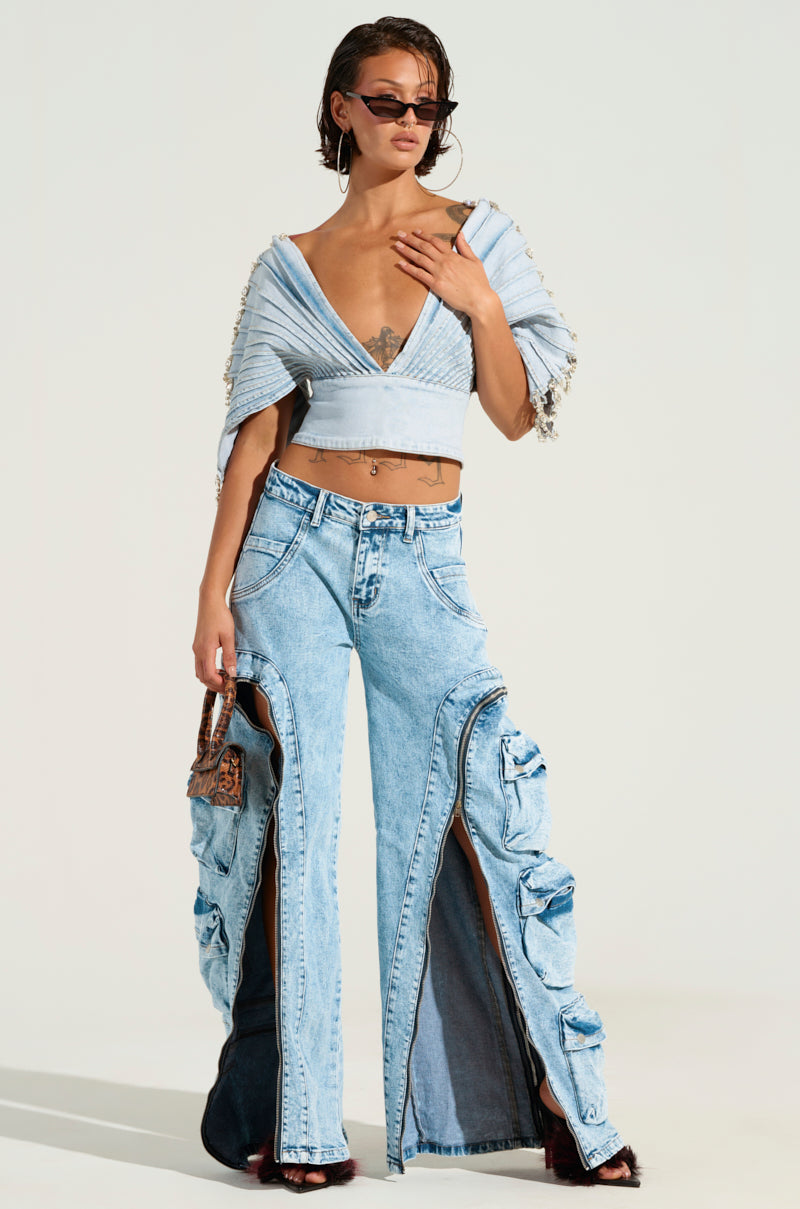 A person dressed in a stylish ensemble pairs a light blue cropped top with the distinctive POCKETFUL ZIPPER DENIM JEANS, which feature unique high-rise cuts and ruffled accents. They accessorize with a small bag and sunglasses, posing against a plain backdrop.