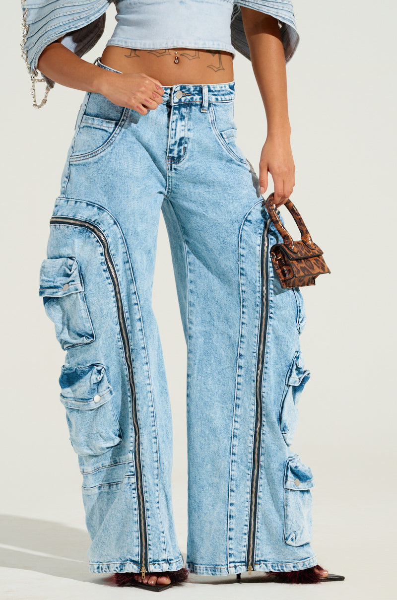 A person is wearing POCKETFUL ZIPPER DENIM JEANS in light blue acid-wash with multiple cargo pockets and holding a small brown patterned handbag. They stand casually, with part of a blue top visible.