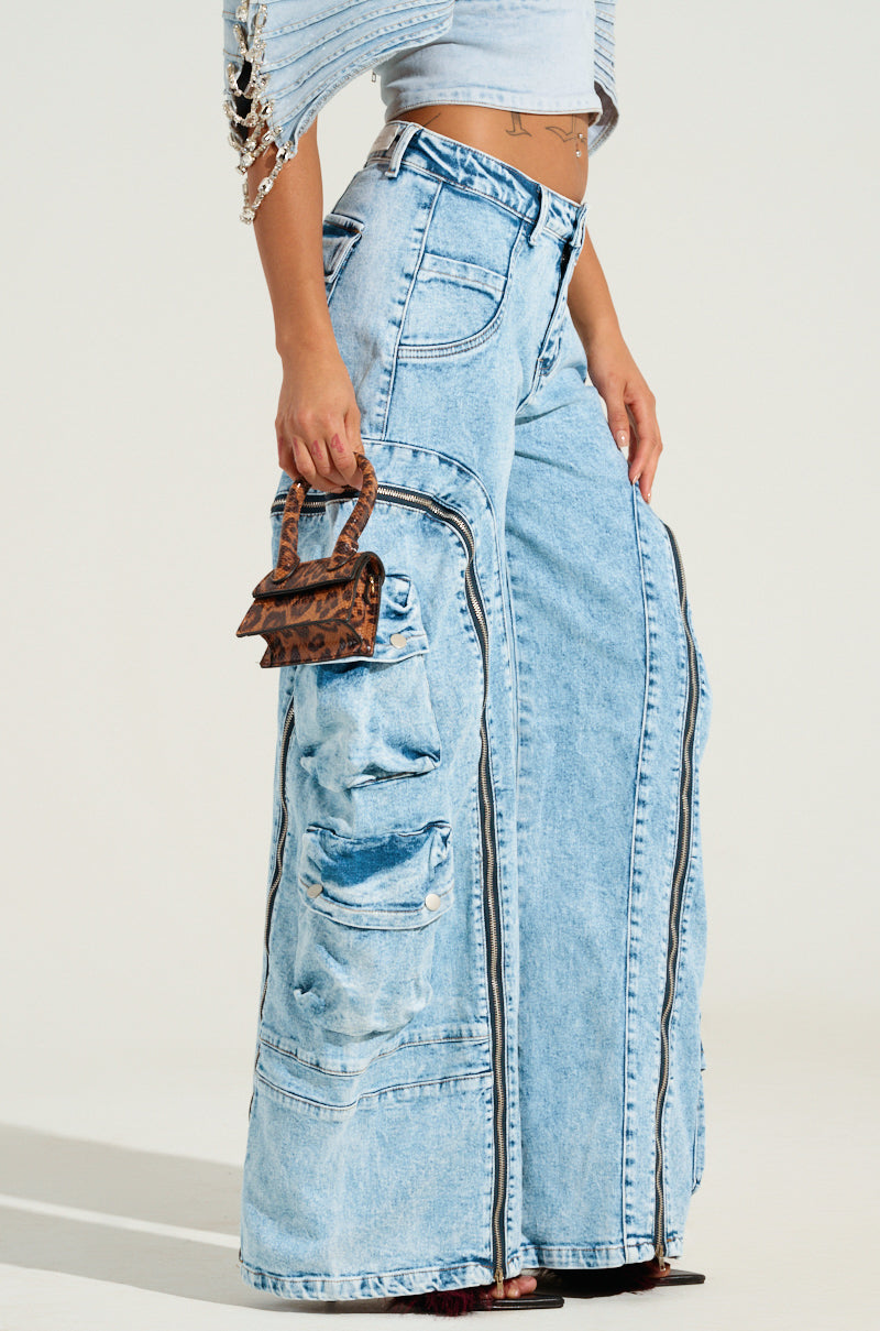 A person wearing the POCKETFUL ZIPPER DENIM JEANS in light blue, featuring a high rise and wide leg design with cargo pockets and zippers, is carrying a small brown patterned handbag. They are also sporting a light-colored top with dangling embellishments on one sleeve.