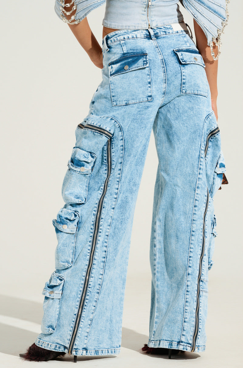 A person wearing the POCKETFUL ZIPPER DENIM JEANS, characterized by their high-rise design, light blue denim color, multiple pockets, and sleek black zippers, stands against a plain background. The jeans offer a loose fit with the person’s hand casually resting on their hip.
