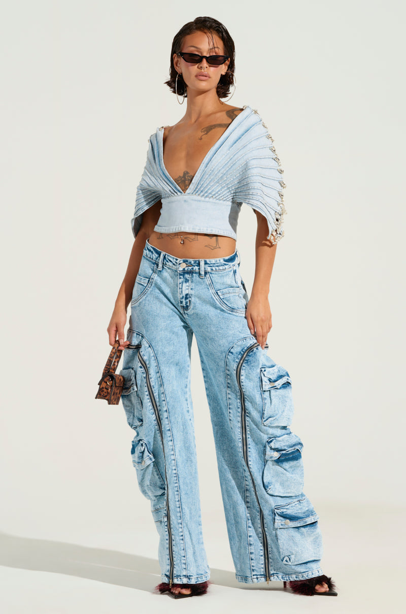 A person stands confidently, styled in an eye-catching ensemble featuring the POCKETFUL ZIPPER DENIM JEANS—high-waisted with unique gathered details. They pair it with a light blue cropped top adorned with wide sleeves and embellished accents, complementing the look with a small patterned handbag, sunglasses, and earrings.