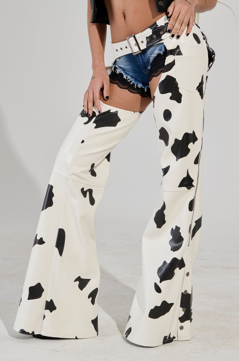 A person wearing the MOO OVER Cow Print Faux Leather Chaps and denim shorts paired with a white belt and a black leather jacket, standing against a plain background. Their hands rest confidently on their hips.