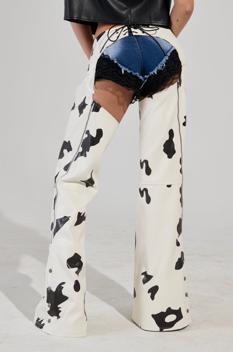 A person is wearing the MOO OVER COW PRINT FAUX LEATHER CHAPS over denim shorts and black lace shorts. They face away, showcasing the unique design with a lace-up back panel. A metallic buckle waist belt adds a sleek touch to the ensemble. The plain gray backdrop enhances the distinct style of the outfit.