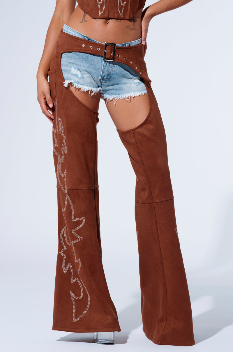 A person dressed in the Smoke Hour Suede Chaps in brown over denim shorts, featuring embroidered designs and a belt at the waist. The look is completed with transparent high heels against a plain white background.