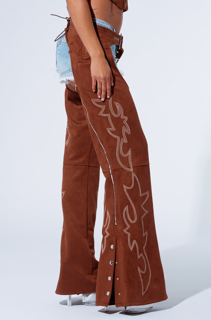 A person is wearing the SMOKE HOUR SUEDE CHAPS IN BROWN from AKIRA Label, which showcase Western-inspired embroidery over denim shorts. The chaps feature intricate white stitching and silver snaps at the bottom. Complementing the look are gray high-heeled shoes against a plain white background.