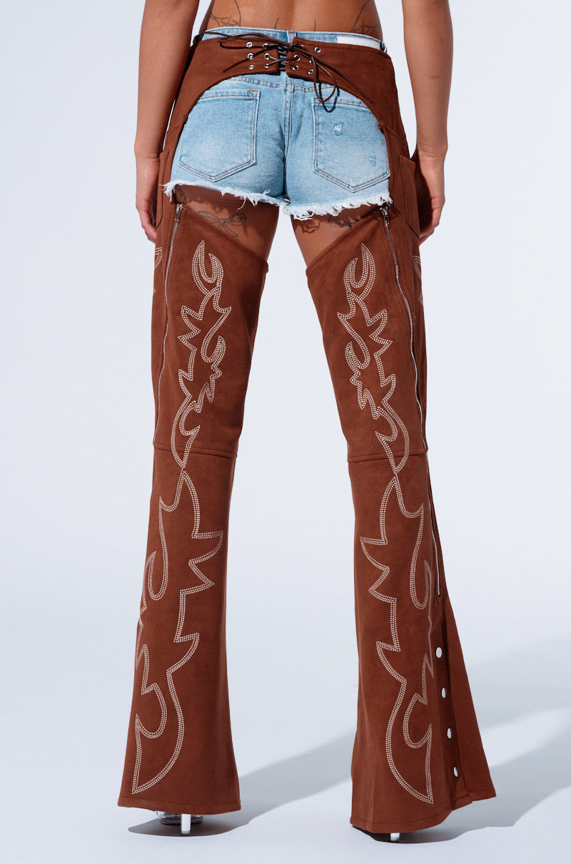 Rear view of a person wearing light blue frayed denim shorts paired with the SMOKE HOUR SUEDE CHAPS IN BROWN from AKIRA Label, featuring intricate white stitching designs. The Western-inspired chaps are secured with lacing at the waistband and extend to the ankles, flaring out slightly at the bottom.