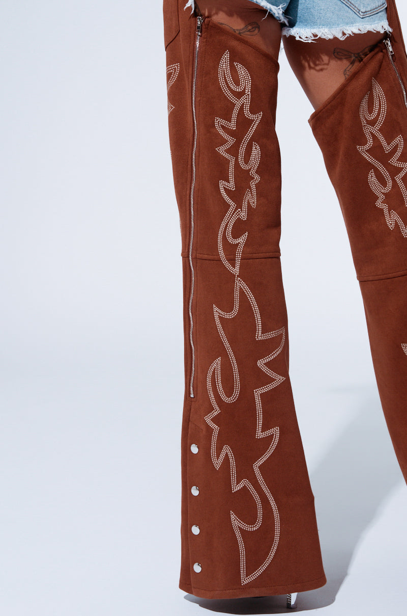 Partial view of a person from the waist down wearing the "SMOKE HOUR SUEDE CHAPS IN BROWN," over-the-knee boots with intricate, white embroidered patterns. The Western-inspired boots feature silver buttons and zippers on the sides, and the person is also wearing frayed denim shorts from the AKIRA Label.
