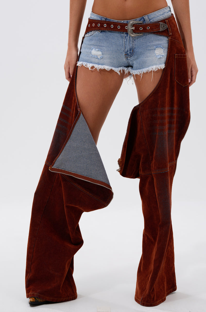 A person stands against a plain white background wearing frayed denim shorts, a belt, and INCREDIBLE VELVET DENIM CHAPS—one leg fully on, the other revealing the shorts.