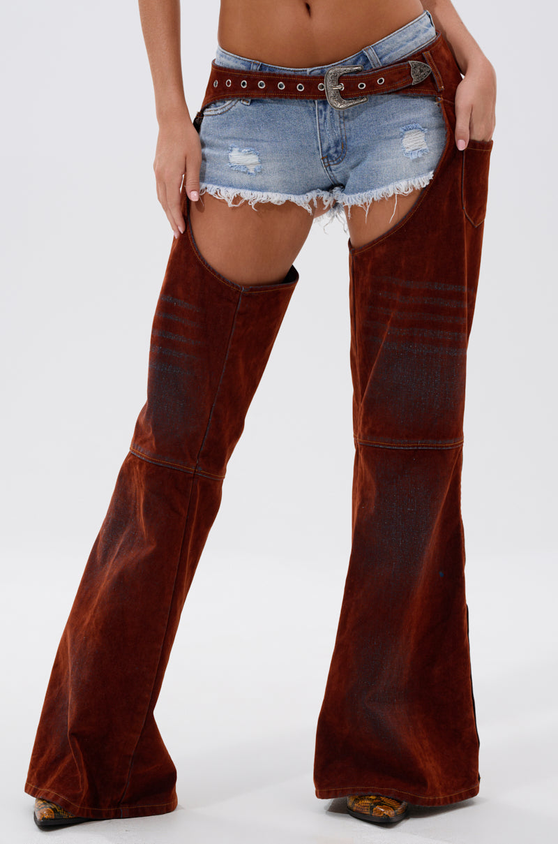 A person models INCREDIBLE VELVET DENIM CHAPS over cowboy boots, paired with distressed shorts and a wide belt, embodying modern cowgirl couture against a plain white background.